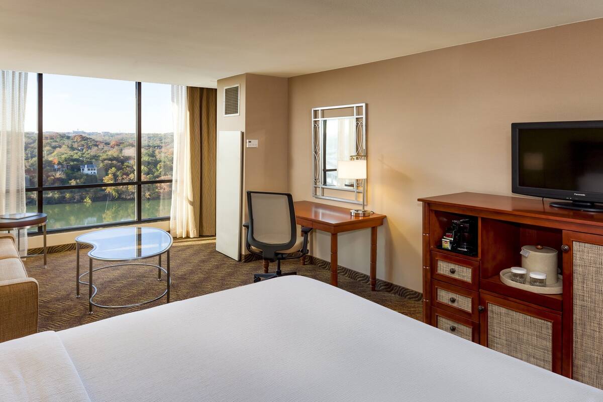 A hotel room with a large bed, a desk with a chair, a television, and a view of a cityscape through large windows.