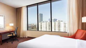 Une chambre d’hôtel dotée d’une grande fenêtre, d’un lit, d’un bureau avec une chaise et offrant une vue sur le paysage urbain.