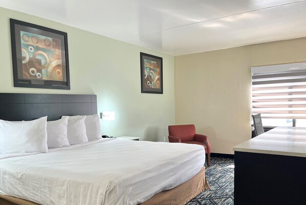 Habitación de hotel con una cama grande, un escritorio, dos cuadros abstractos enmarcados, una silla roja y una ventana con persianas.