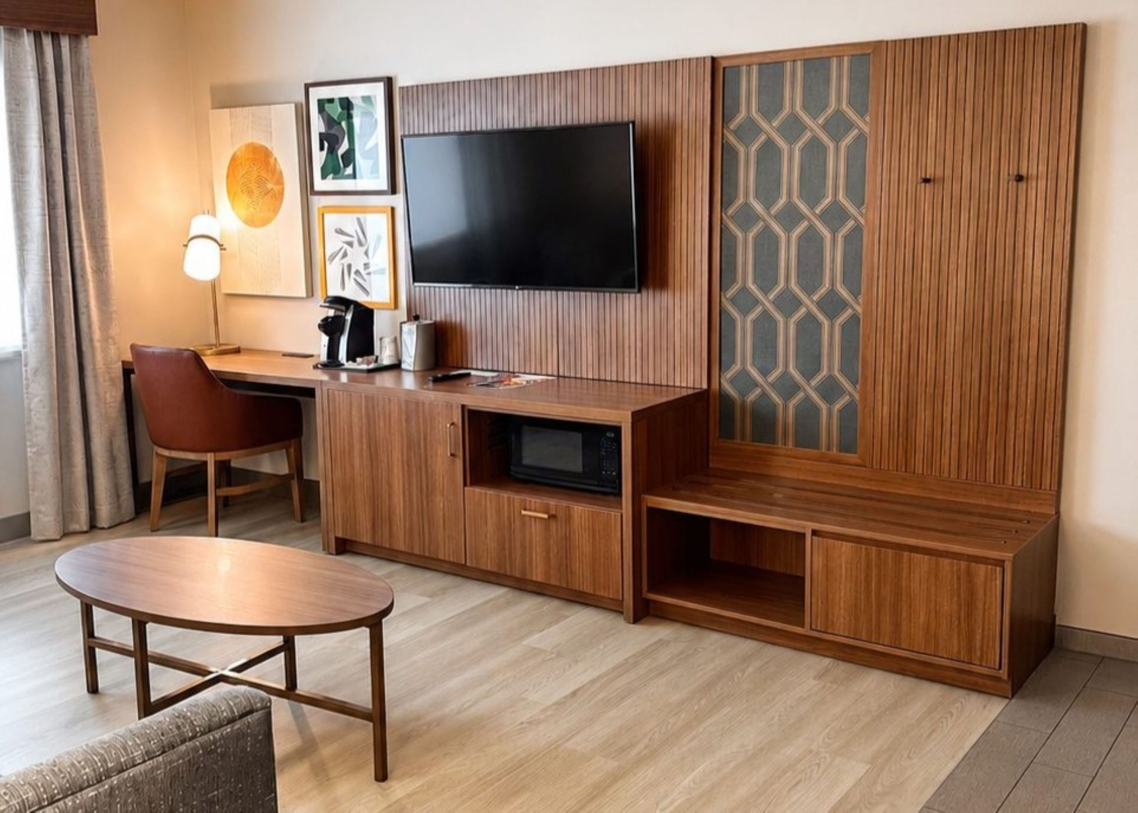A modern hotel room with a wooden entertainment unit, a flat-screen TV, a desk, and a seating area with a round table.
