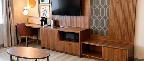 A modern hotel room with a wooden entertainment unit, a flat-screen TV, a desk, and a seating area with a round table.