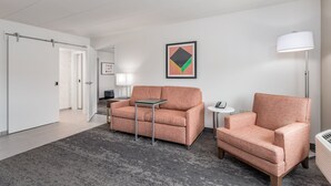 Une chambre d’hôtel moderne avec un tapis gris, un canapé, un fauteuil, une petite table, une lampe et un tableau encadré au mur.