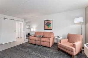 A modern hotel room with a grey carpet, a sofa, a chair, a small table, a lamp, and a framed picture on the wall. - Holiday Inn Columbus by IHG (Columbus)
