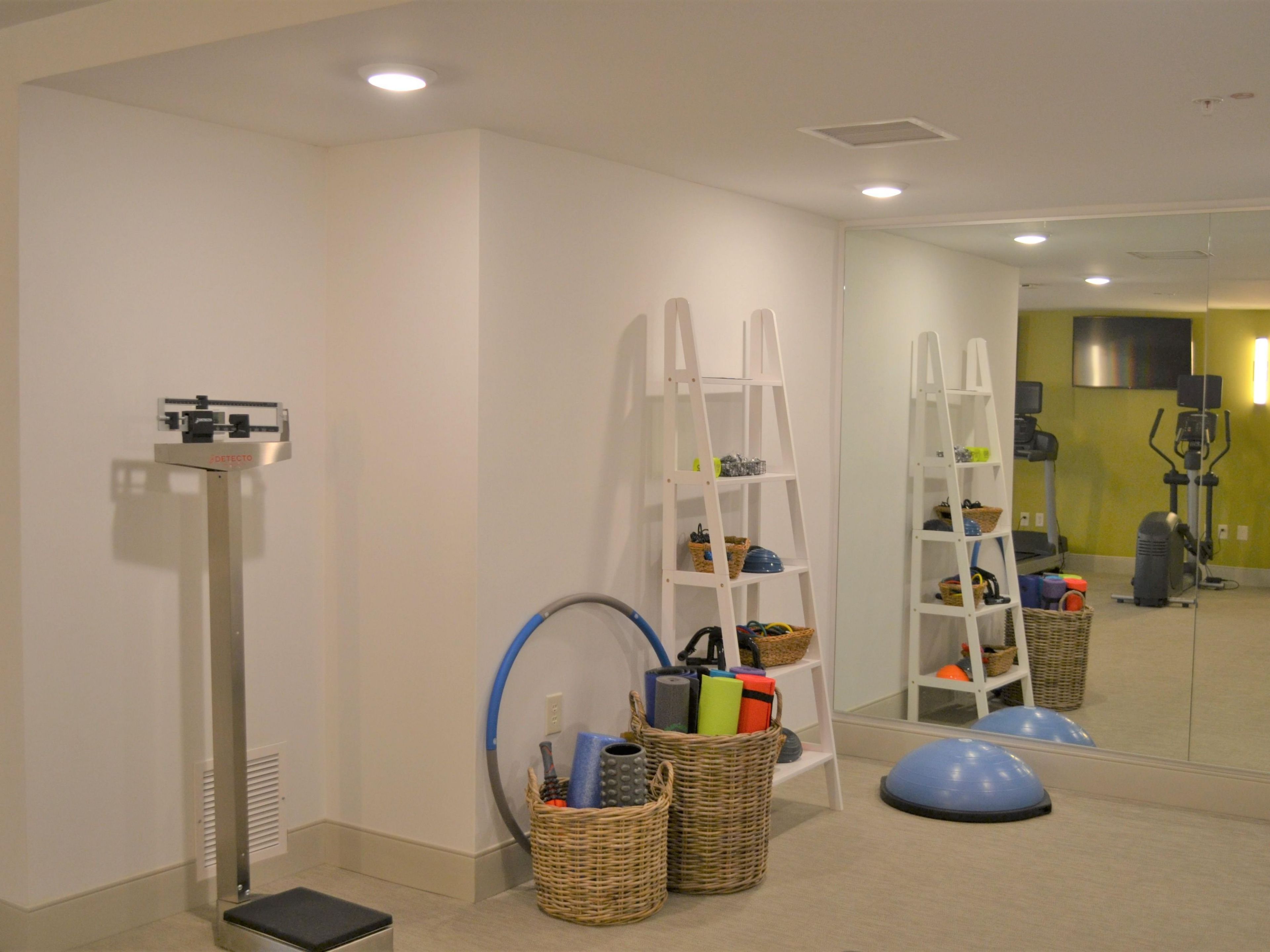 Una habitación con una máquina de ejercicio vertical, dos estanterías blancas con cestas, una pelota de ejercicio azul y un espejo que refleja equipo de gimnasia.