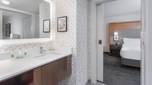 Une salle de bain d’hôtel moderne, dotée d’un grand miroir, de deux lavabos et d’une coiffeuse. Un tableau encadré orne le mur, et une table de chevet avec une lampe se trouve à côté du lit.