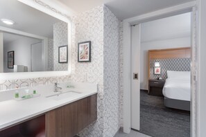 A modern hotel bathroom with a large mirror, two sinks, and a vanity. There is a framed picture on the wall and a nightstand with a lamp next to the bed. - Holiday Inn Columbus by IHG (Columbus)