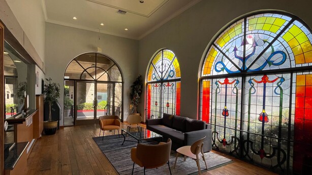 A spacious lobby with wooden flooring, a black sofa, and two armchairs. Large stained glass windows with colorful designs are visible. - Portside Inn & Suites (San Pedro)