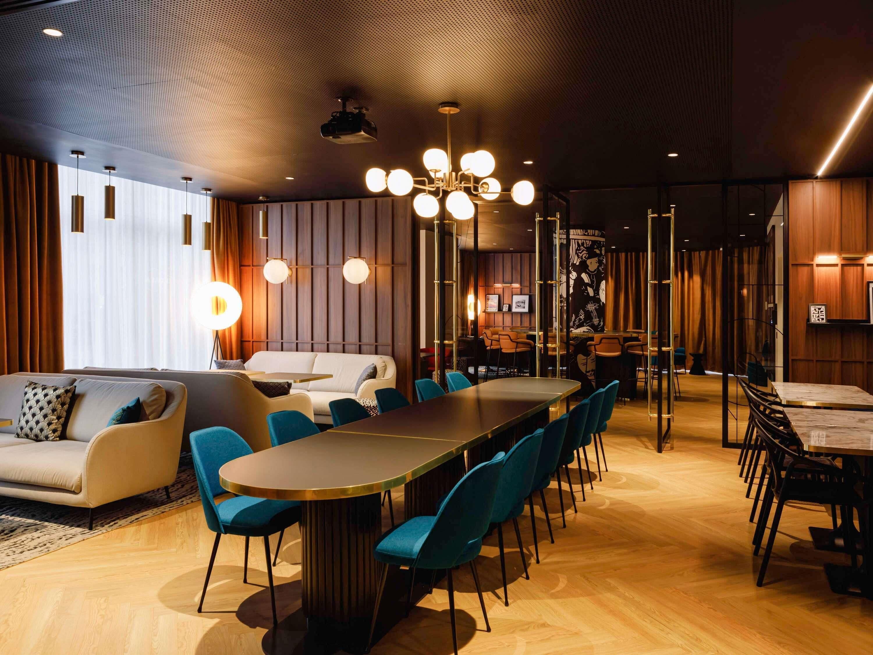 A modern hotel lobby with a long dining table, blue chairs, a sofa, and a ceiling projector.