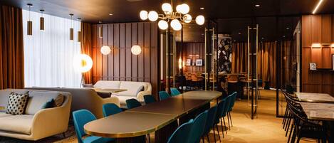 A modern hotel lobby with a long dining table, blue chairs, a sofa, and a ceiling projector.