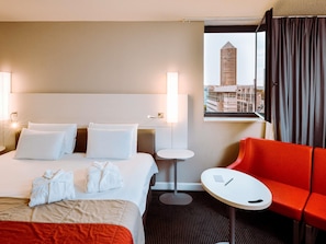A hotel room with a bed, two pillows, a red chair, a small table, and a view of a cityscape. - Mercure Lyon Centre Saxe Lafayette (Lyon)
