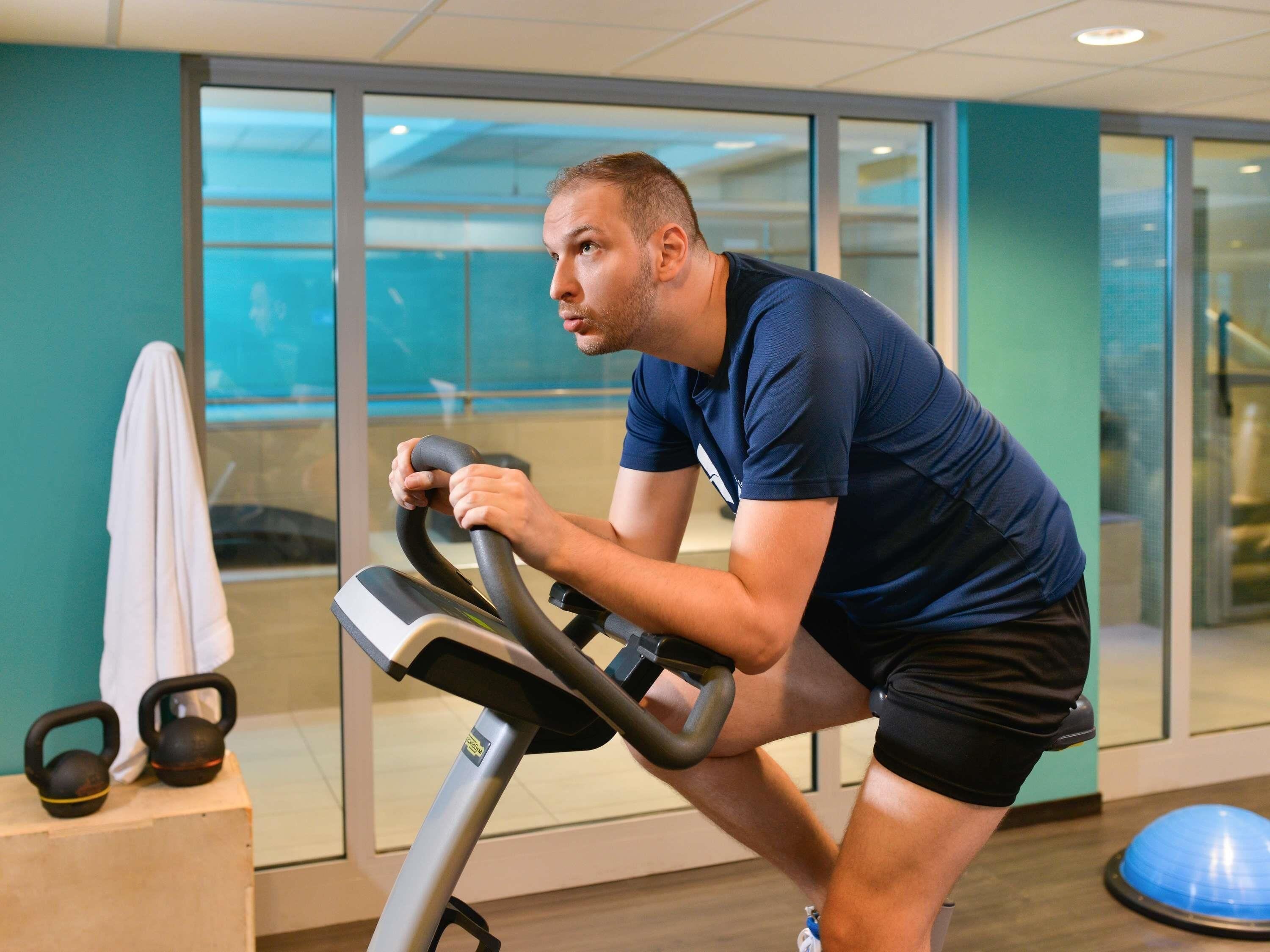Een man die op een hometrainer aan het sporten is in een sportschool, met een handdoek en kettlebells op een aanrecht, en een blauwe fitnessbal op de achtergrond.