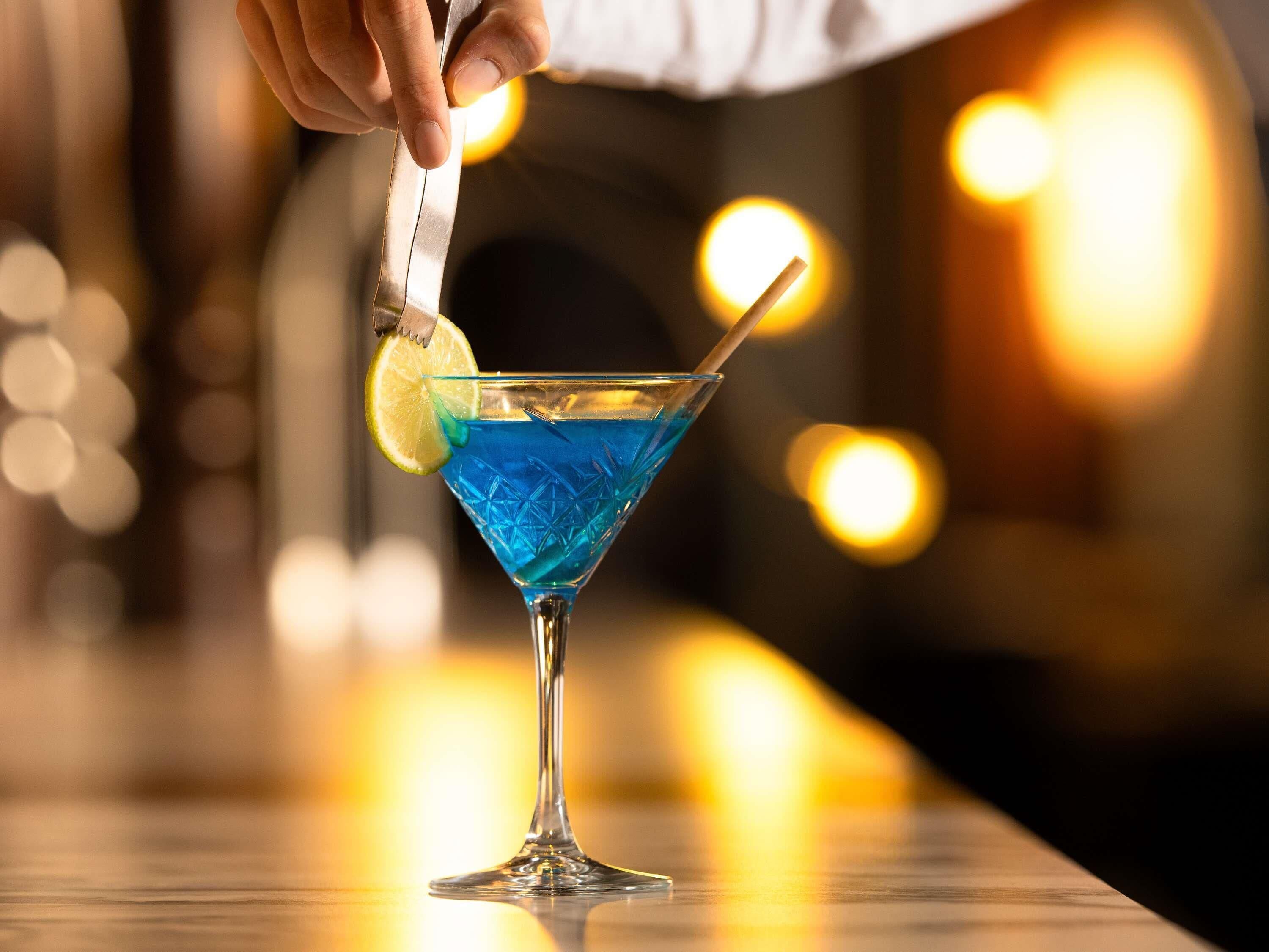 A martini glass with a blue liquid, a lime wedge, and a straw on a bar counter.
