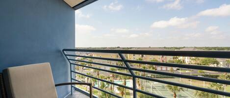 A balcony with a wicker chair, a view of a tennis court, and a residential area in the background.