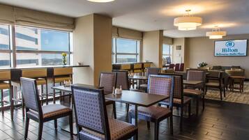 Comedor de hotel con varias mesas y sillas, ventanas amplias y el letrero Hilton Melbourne Rialto Place a la vista.