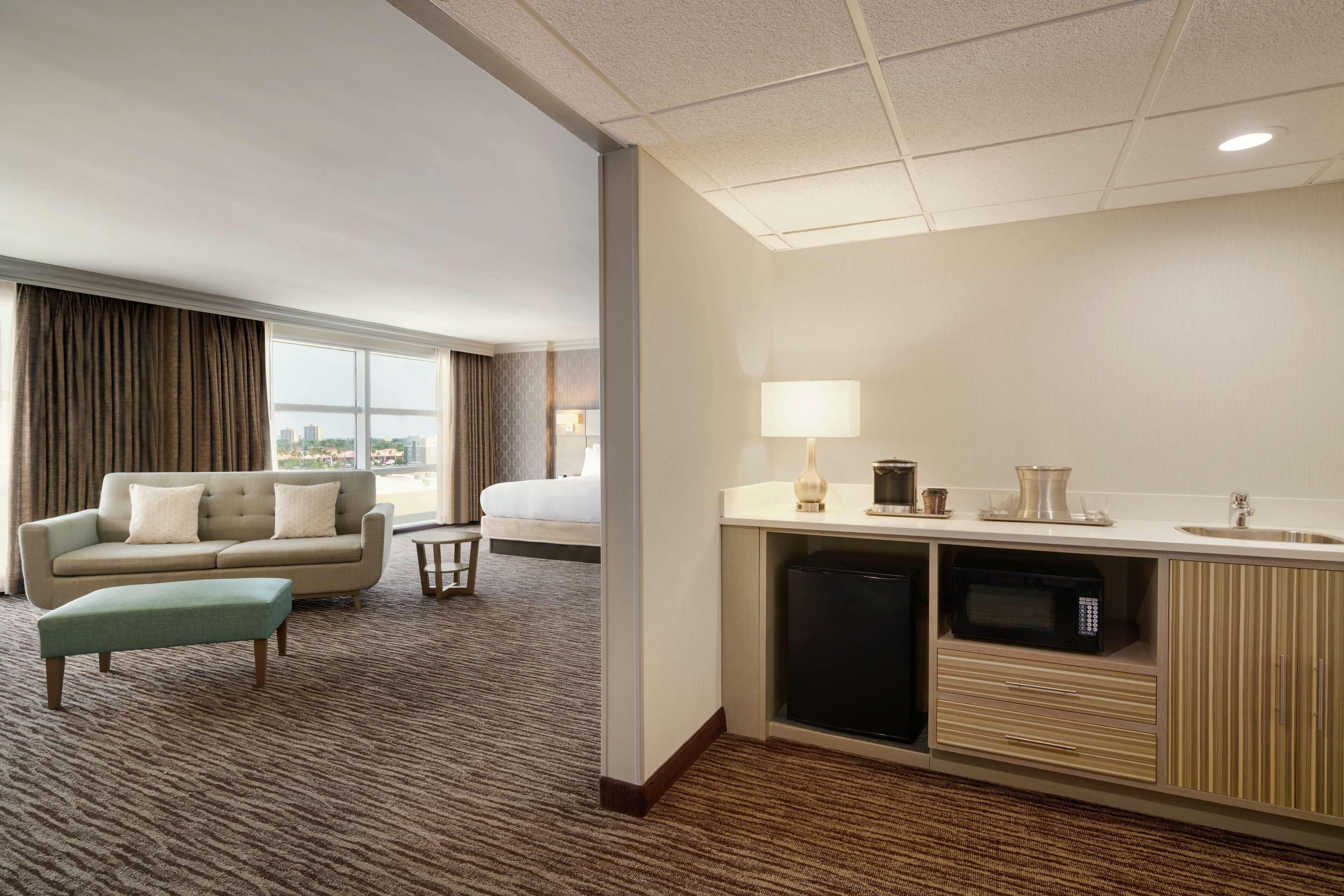 A hotel room with a bed, a sofa, a green ottoman, a kitchenette with a microwave, a sink, and a window with curtains.
