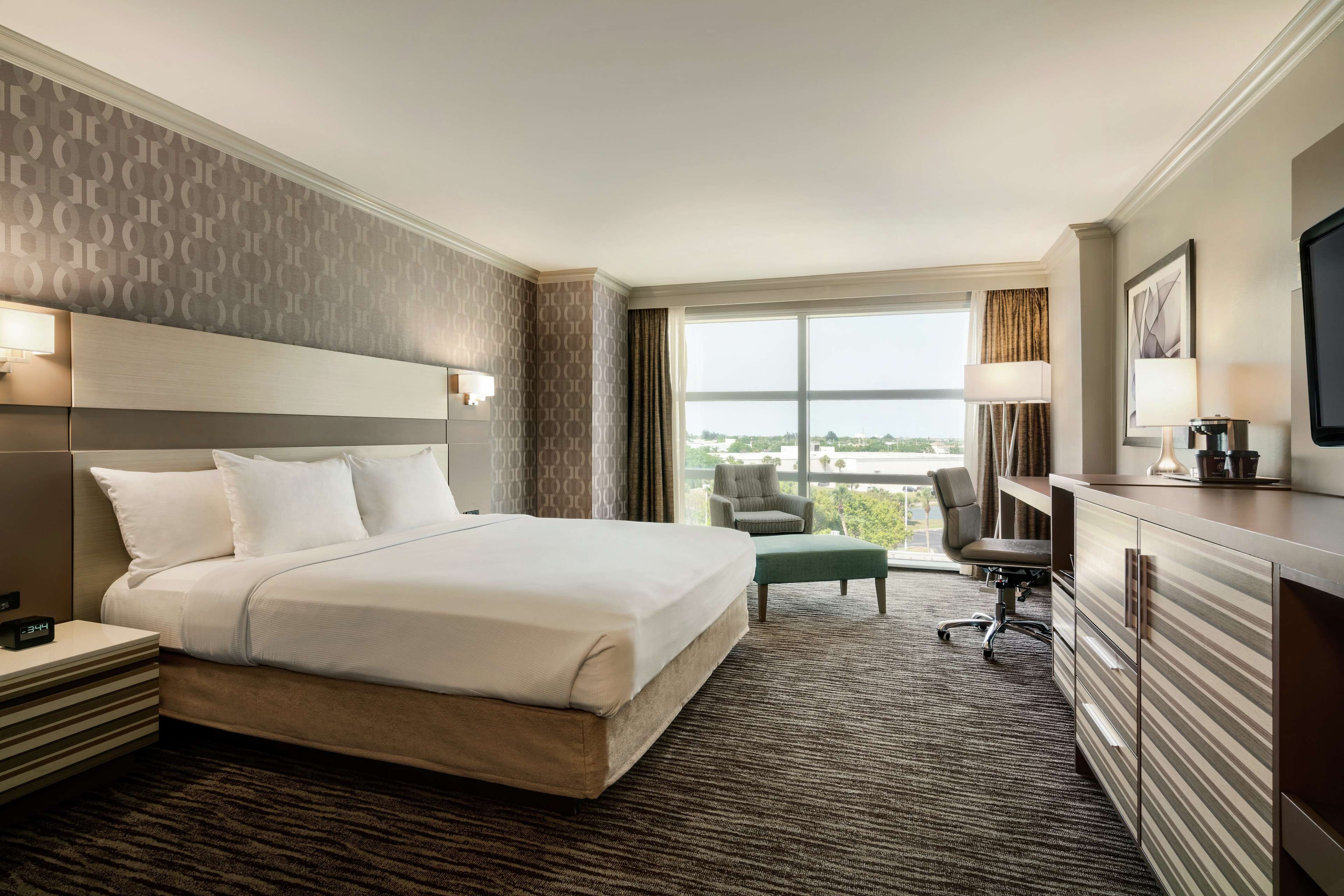 A hotel room with a large bed, a desk, a chair, a television, and a view of the city.