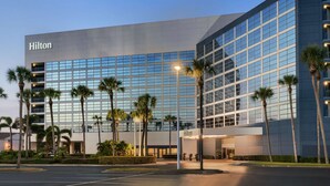 A modern Hilton hotel building with a glass facade, palm trees, and a well-lit entrance. - Hilton Melbourne, FL (Melbourne)