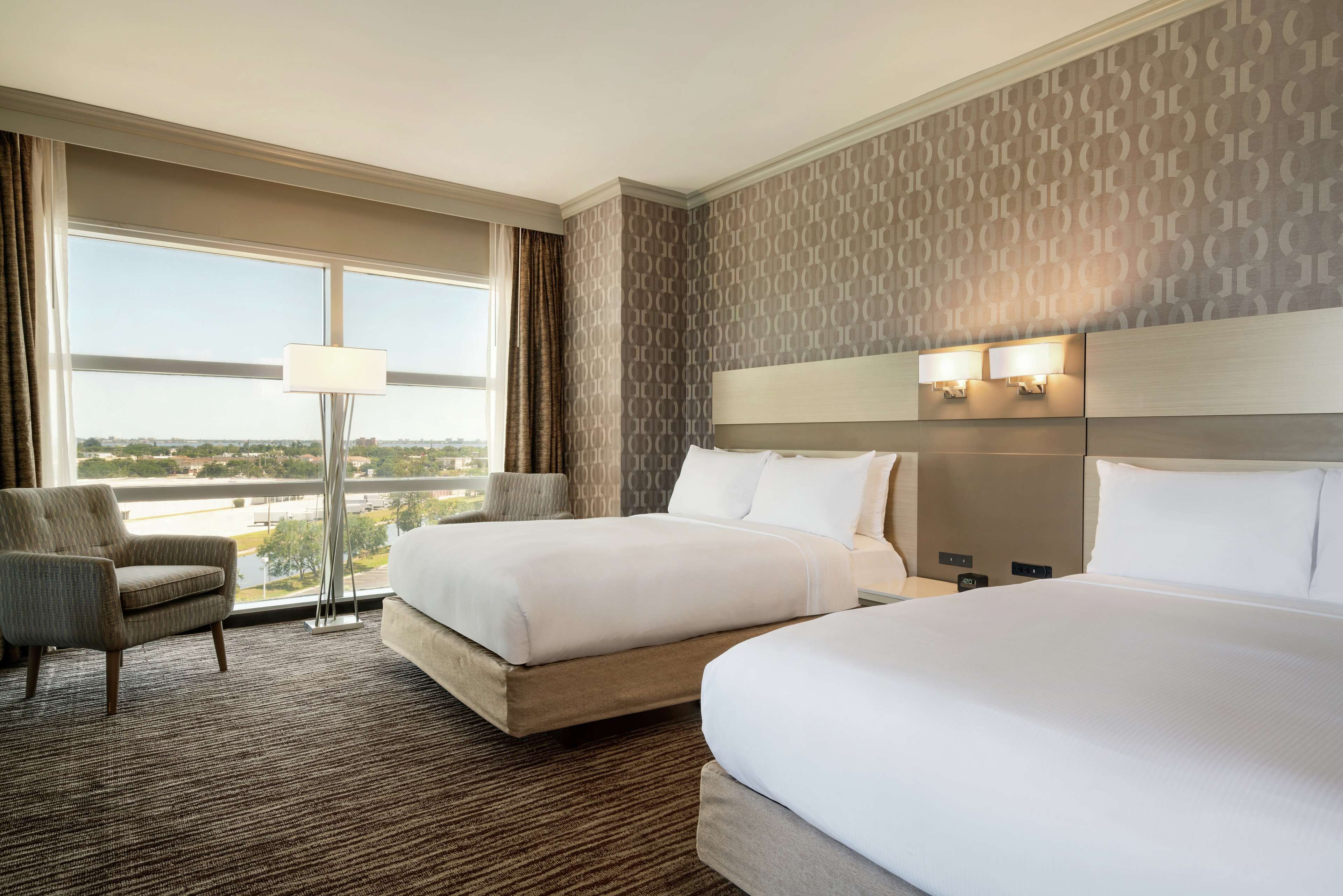 Habitación de hotel con dos camas, una silla, una lámpara y vista a la ciudad.