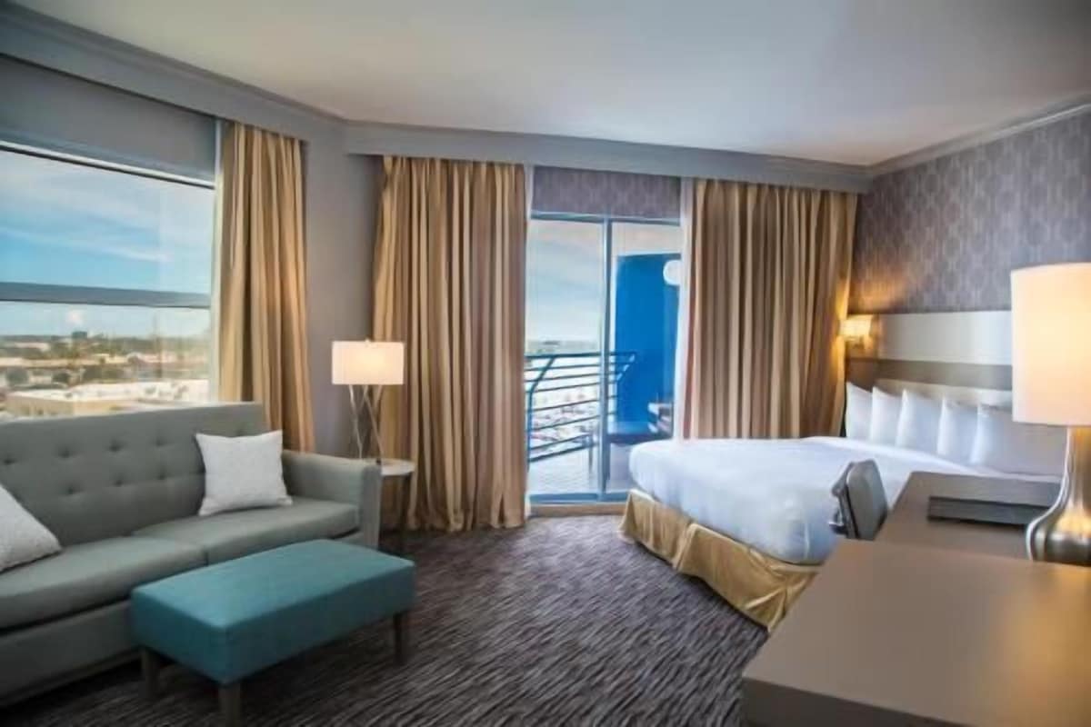 A hotel room with a bed, a sofa, a small table, and a view of the city through large windows.