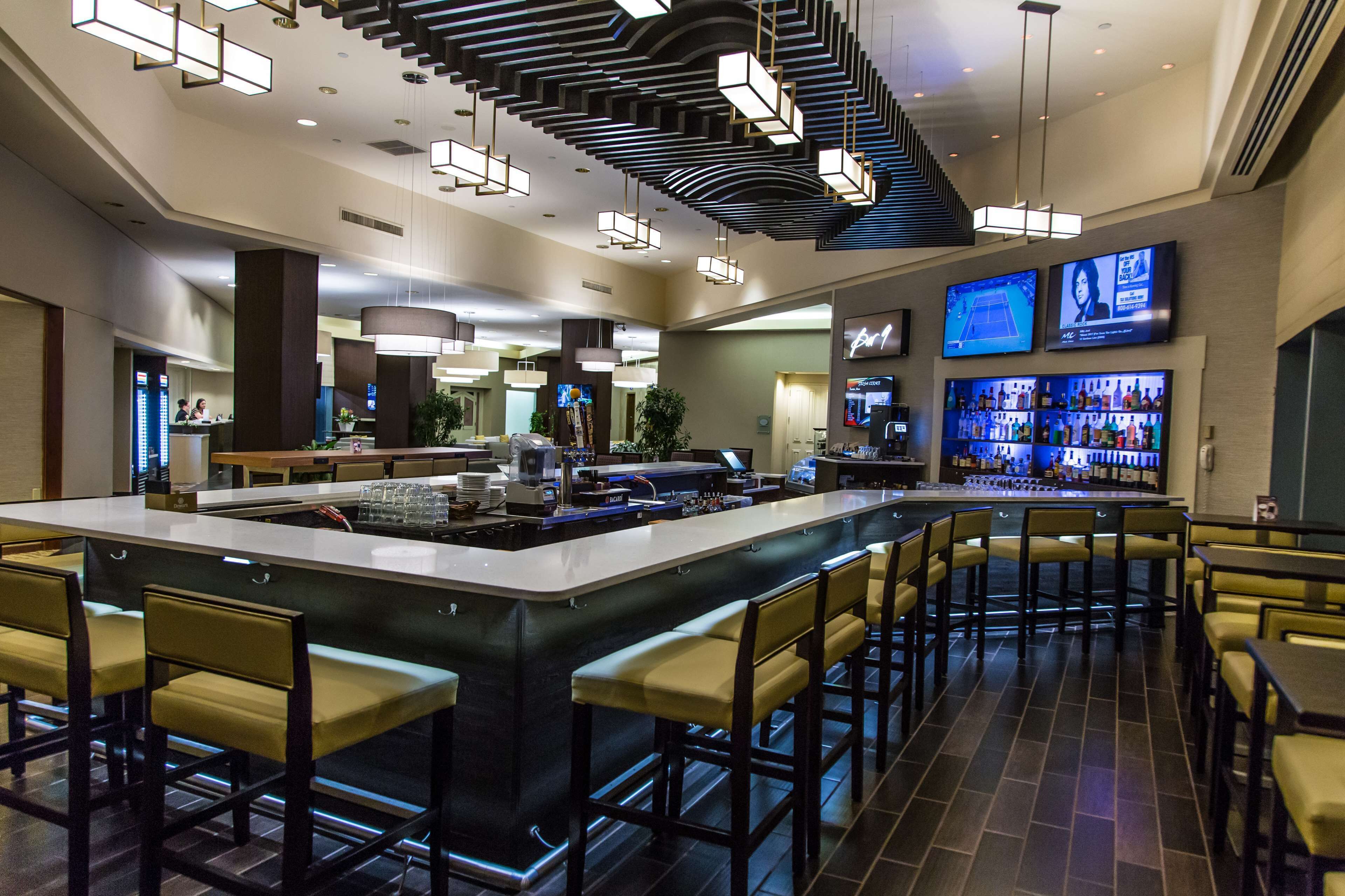 A modern bar area with a central island, bar stools, and a television screen displaying a sports game.