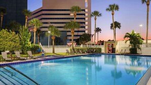 A swimming pool with lounge chairs, palm trees, and a modern building in the background. - Hilton Melbourne, FL (Melbourne)