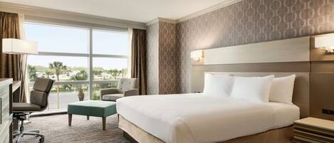 A hotel room with a large bed, a desk with a chair, a small table, and a view of palm trees through the window.