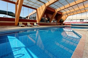 An indoor swimming pool with clear blue water, surrounded by a modern architectural structure with large windows and a roof.