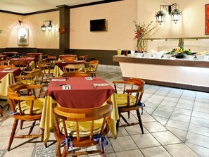 A dining area with multiple tables and chairs, a buffet counter with various food items, and wall-mounted lighting fixtures.