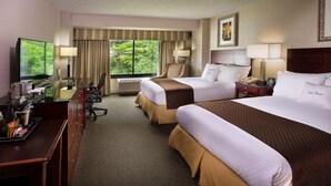 Ein Hotelzimmer mit zwei Betten, einem Schreibtisch mit Fernseher, einem Sessel, einer Schreibtischlampe, einem Telefon und einem Fenster mit Vorhängen.