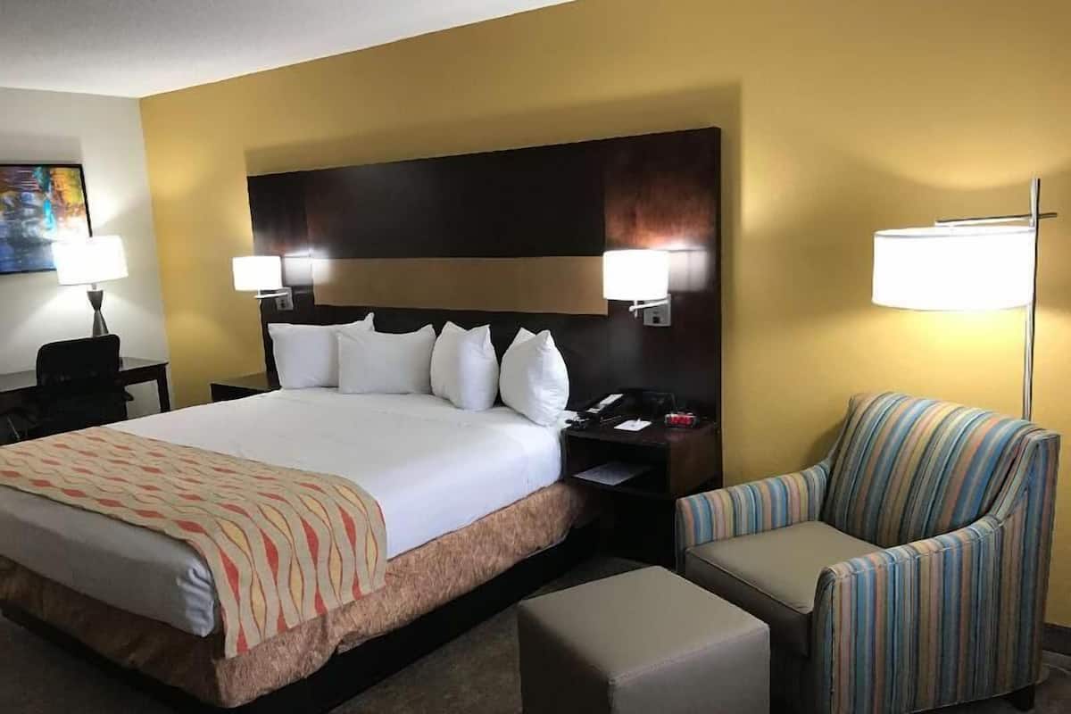 A hotel room with a large bed, two bedside lamps, a striped armchair, a small table, and a painting on the wall.
