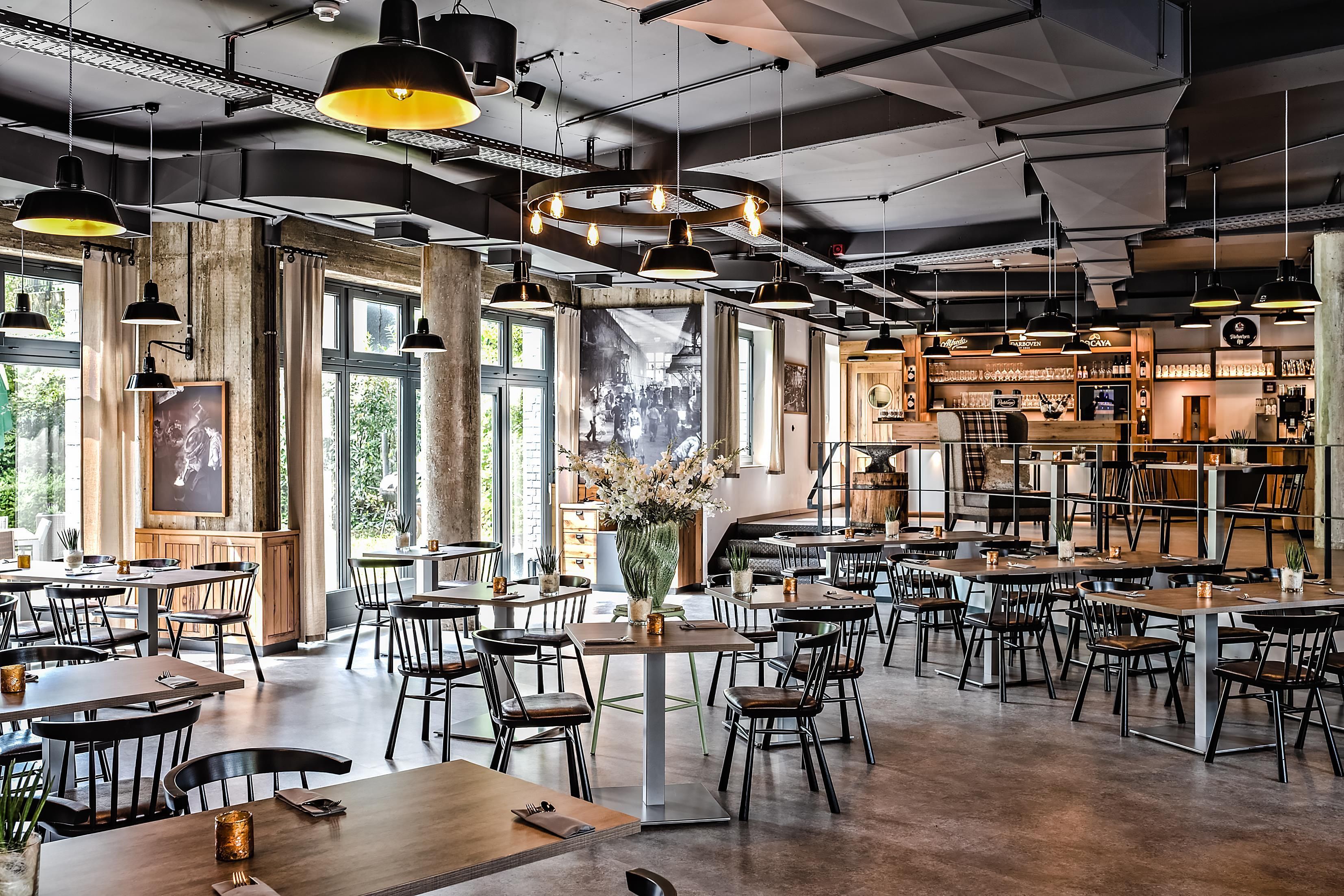 Ein geräumiges Restaurant mit hohen Decken, freiliegenden Balken und großen Fenstern. Tische sind zum Essen gedeckt, und hängende Pendelleuchten sorgen für Beleuchtung.