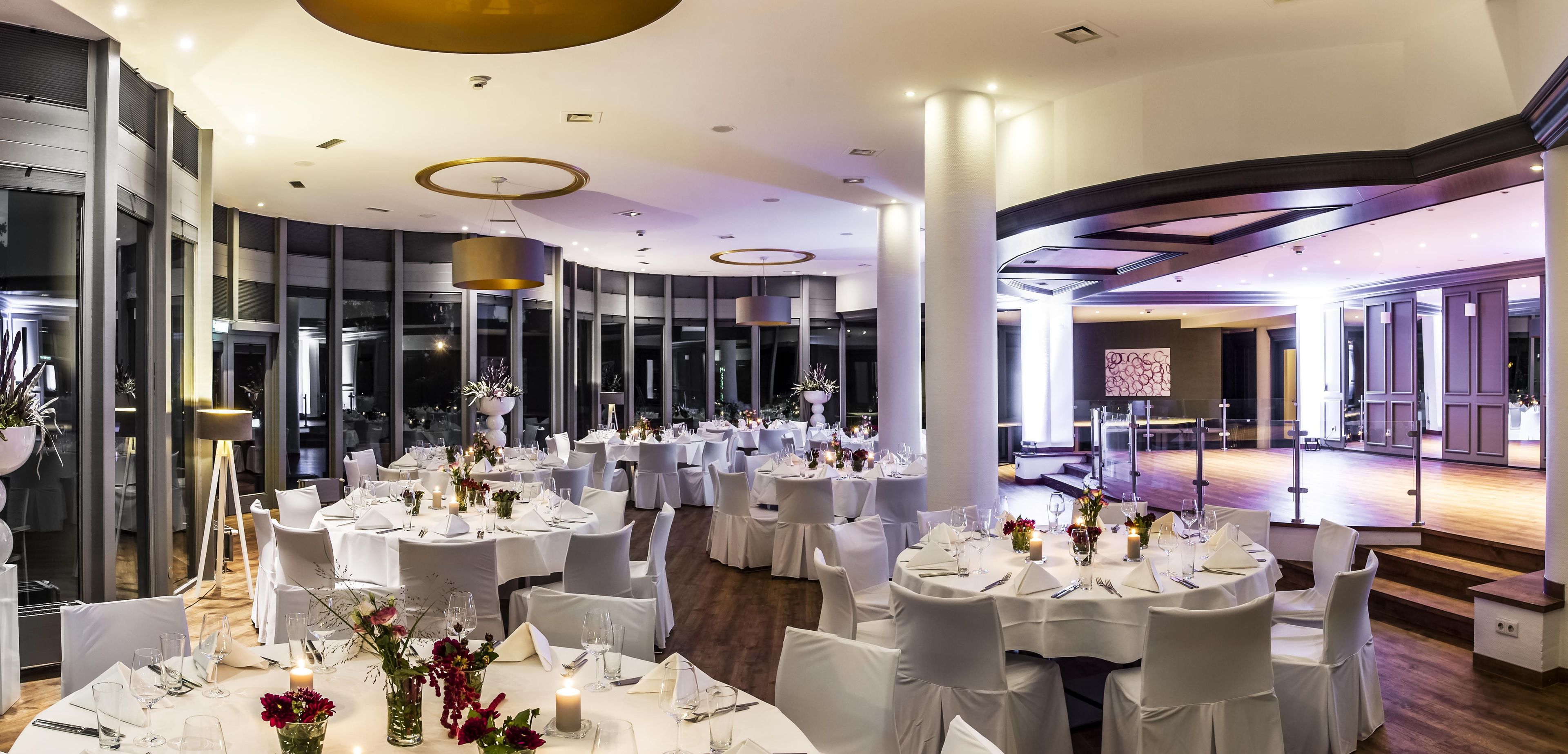 A spacious banquet hall with multiple round tables set for dining, large windows, and a modern ceiling design.