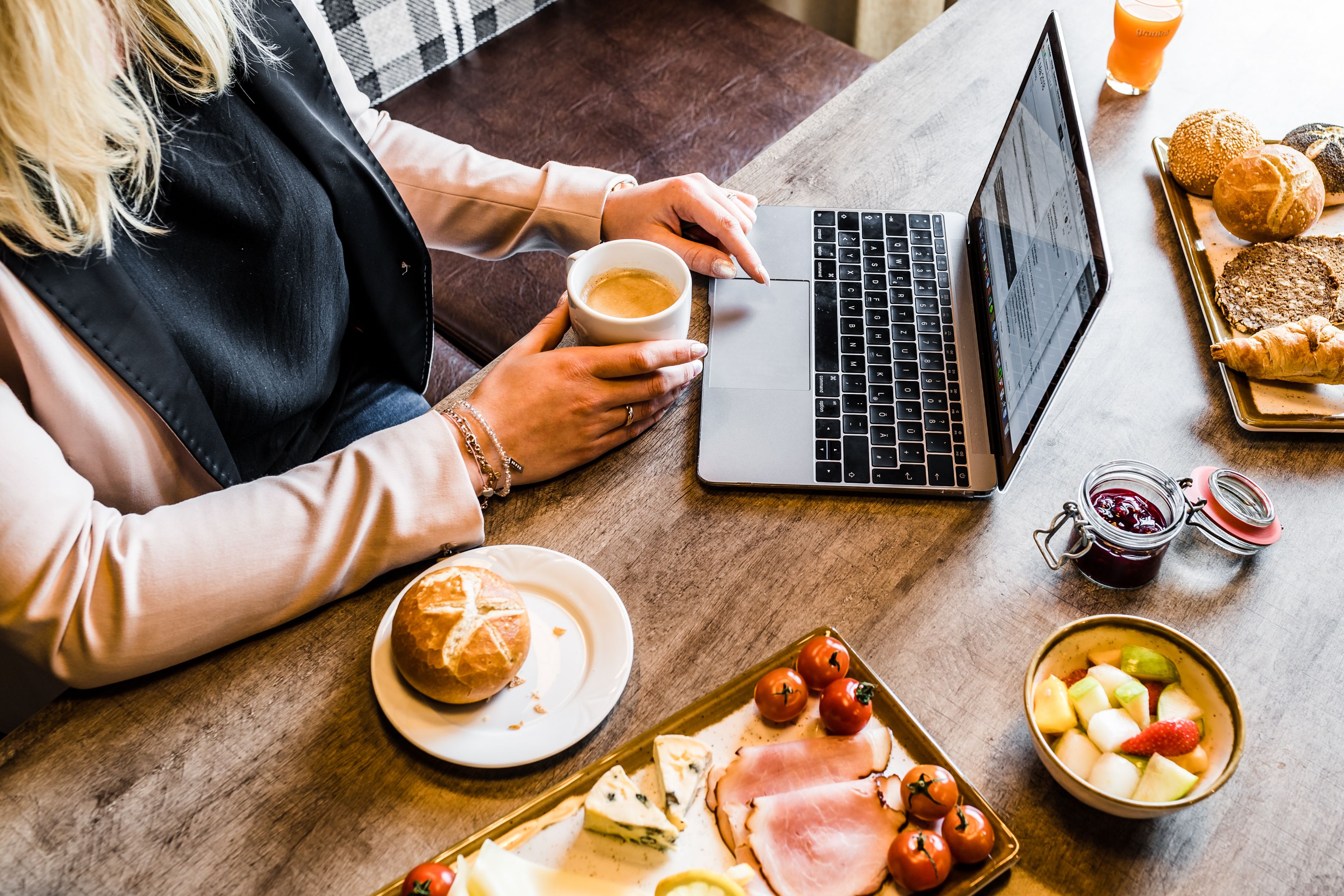 A person using a laptop at a wooden table with food items like bread, cheese, and tomatoes.