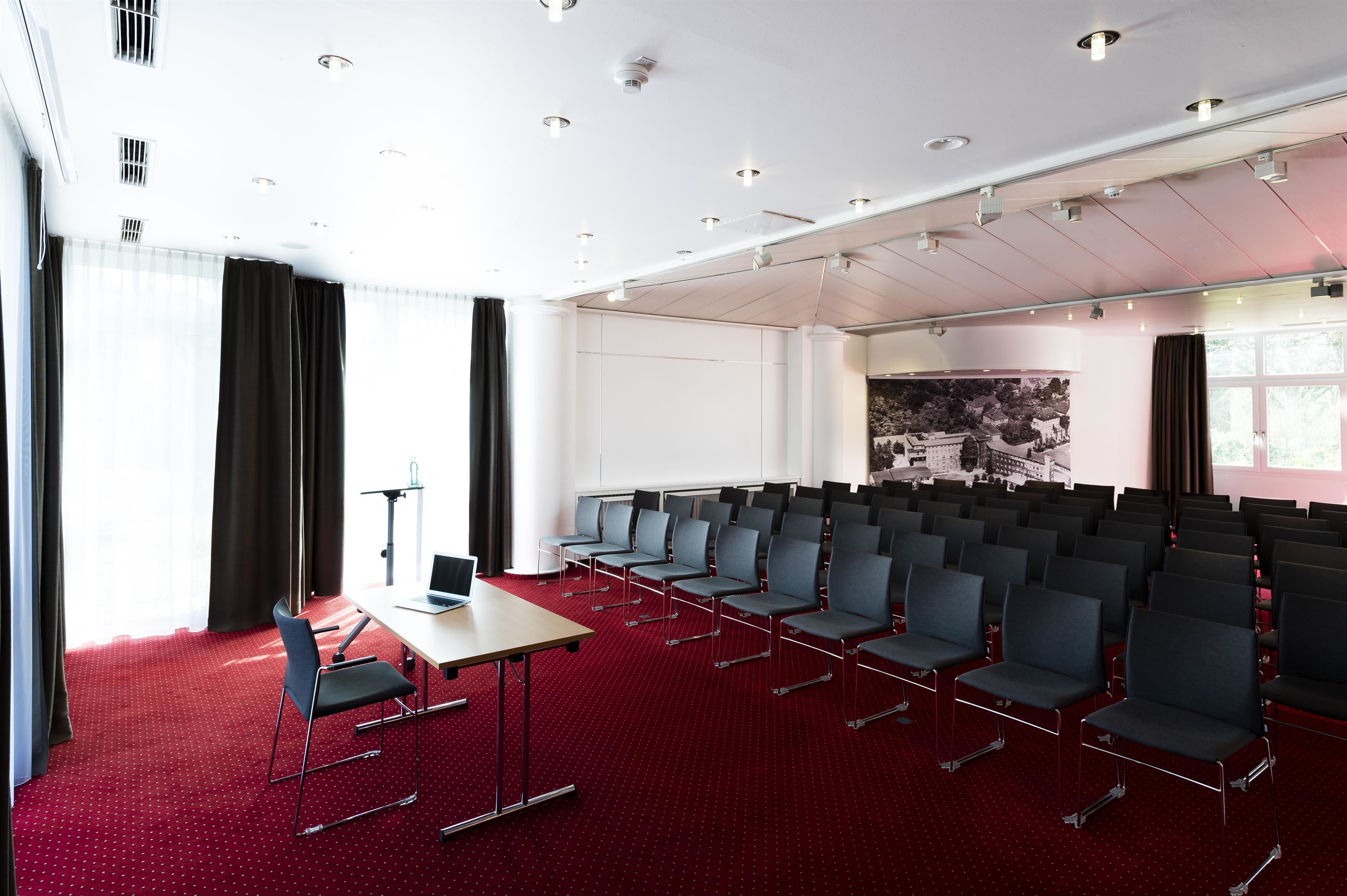 Un salón de conferencias con hileras de sillas, una mesa con una computadora portátil y un ventanal con cortinas.