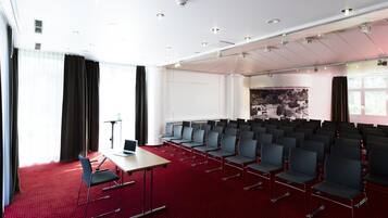 Un salón de conferencias con hileras de sillas, una mesa con una computadora portátil y un ventanal con cortinas.