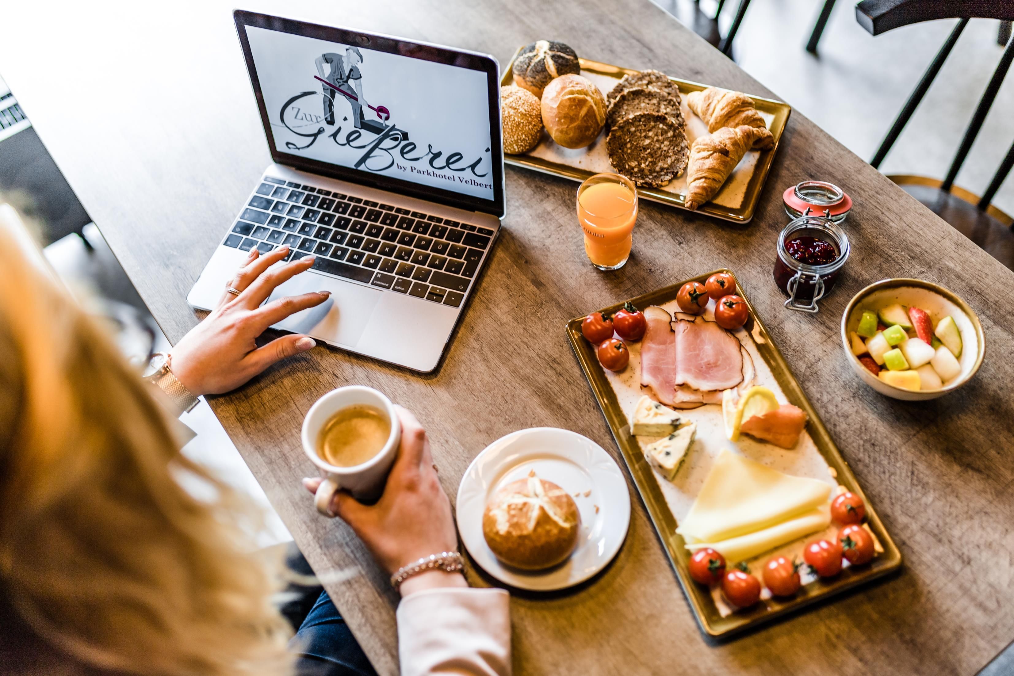 Un ordinateur portable est posé sur une table, à côté d’un plateau de petit-déjeuner contenant des croissants, des tomates, du fromage et du jambon.