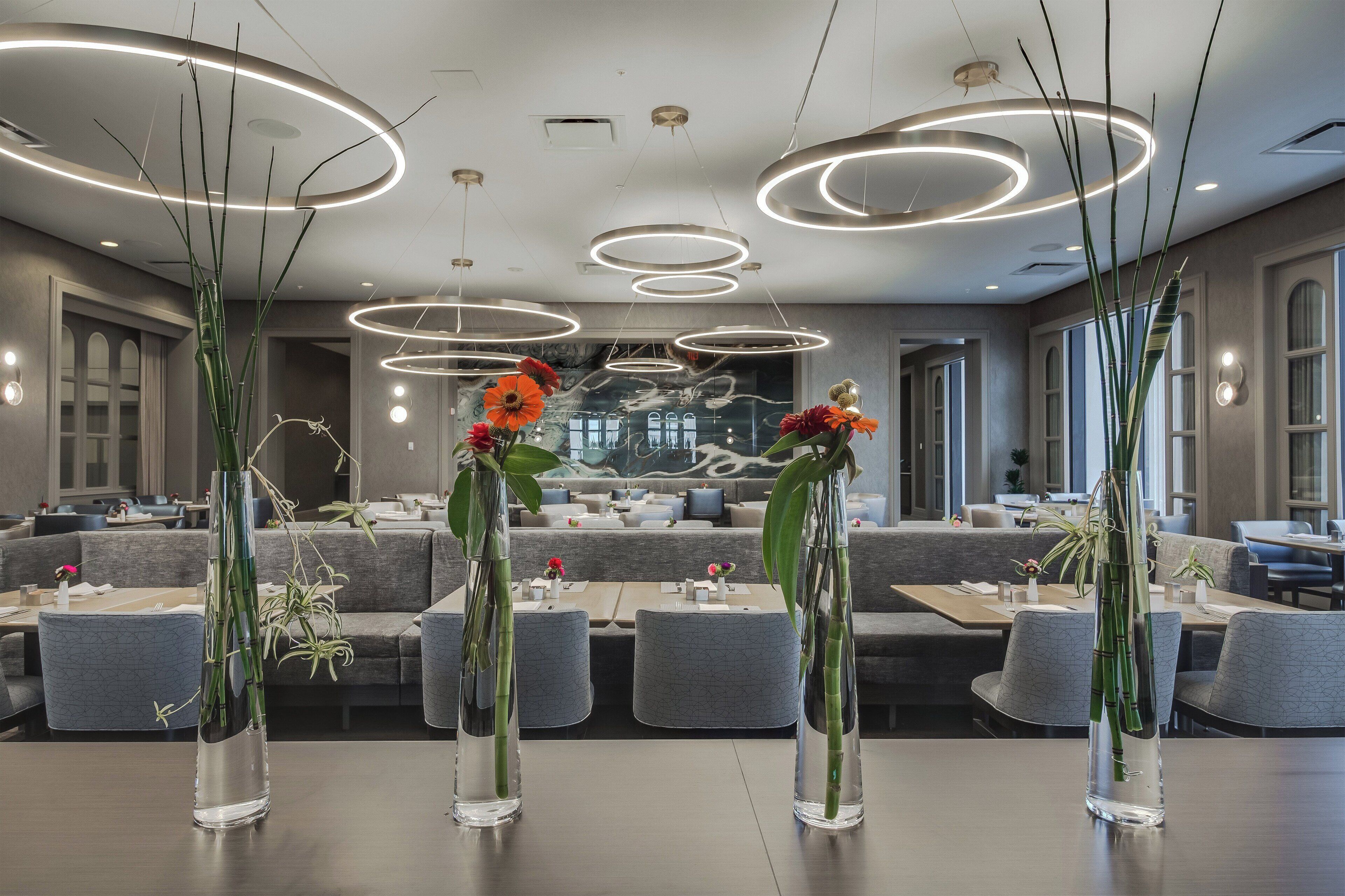 Un comedor moderno con mesas redondas, jarrones transparentes con flores y luces de techo circulares.