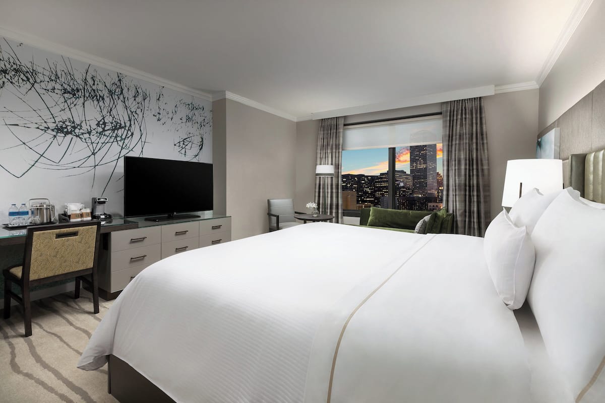 A hotel room with a large bed, a desk with a flat-screen TV, a chair, a coffee maker, and a view of the city at sunset.