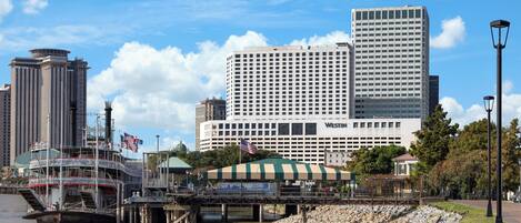 A waterfront hotel with a large building in the background, a dock with a boat, and a pedestrian bridge.
