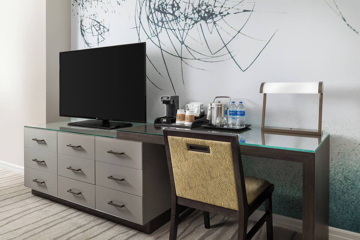 A modern hotel room with a glass desk, a flat-screen TV, a coffee maker, and a chair.