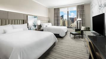 A hotel room with two beds, a desk, a chair, a TV, and a large window with a city view.