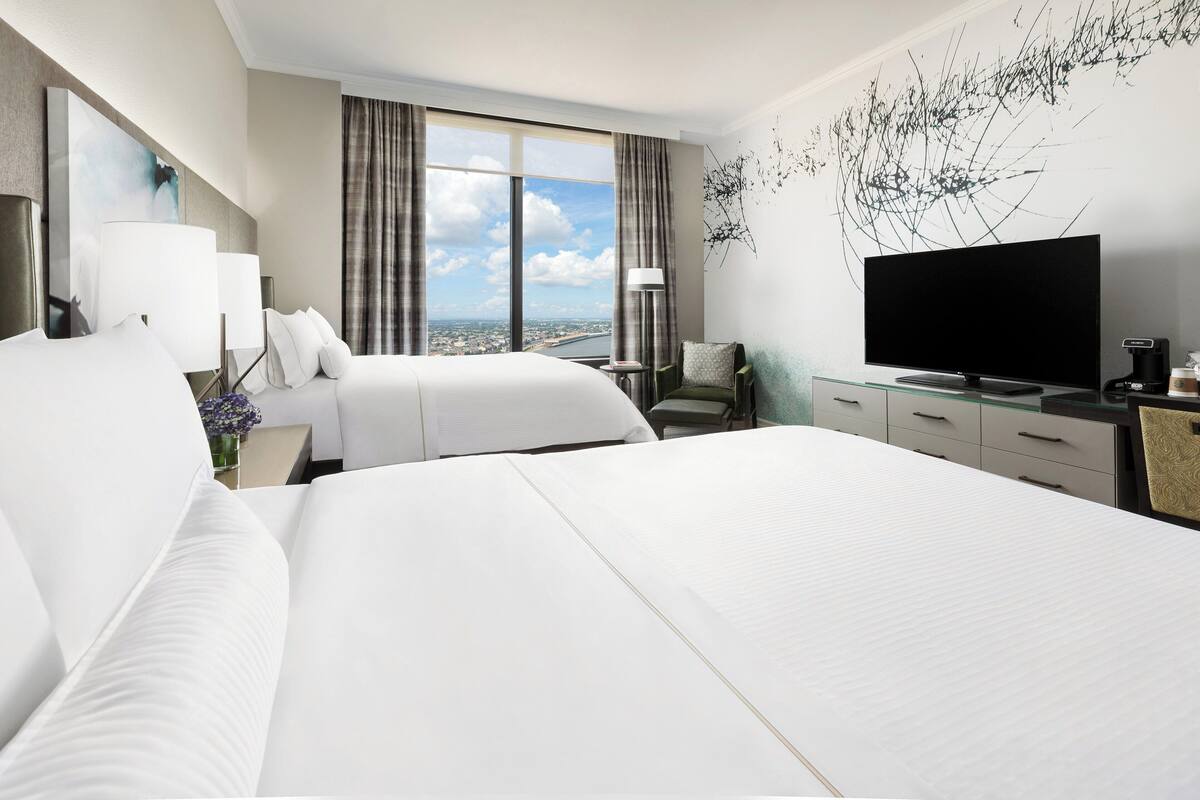 A hotel room with two beds, a large window with a city view, a flat-screen TV, and a desk with a lamp.