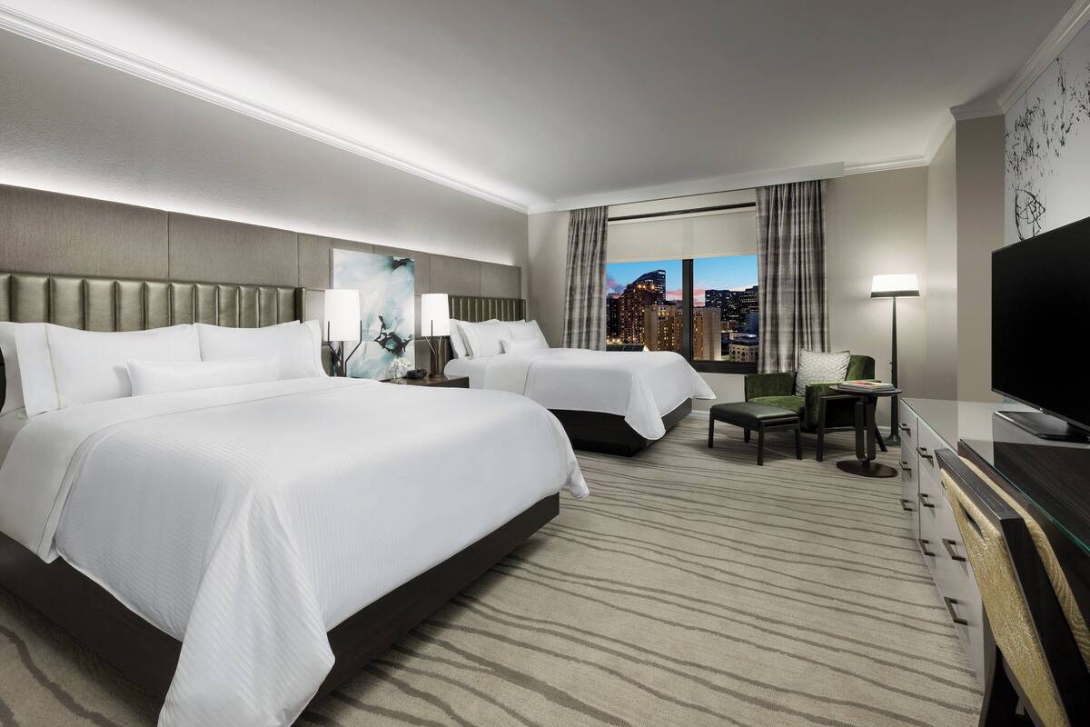 A hotel room with two beds, a TV, a desk, and a view of the cityscape.