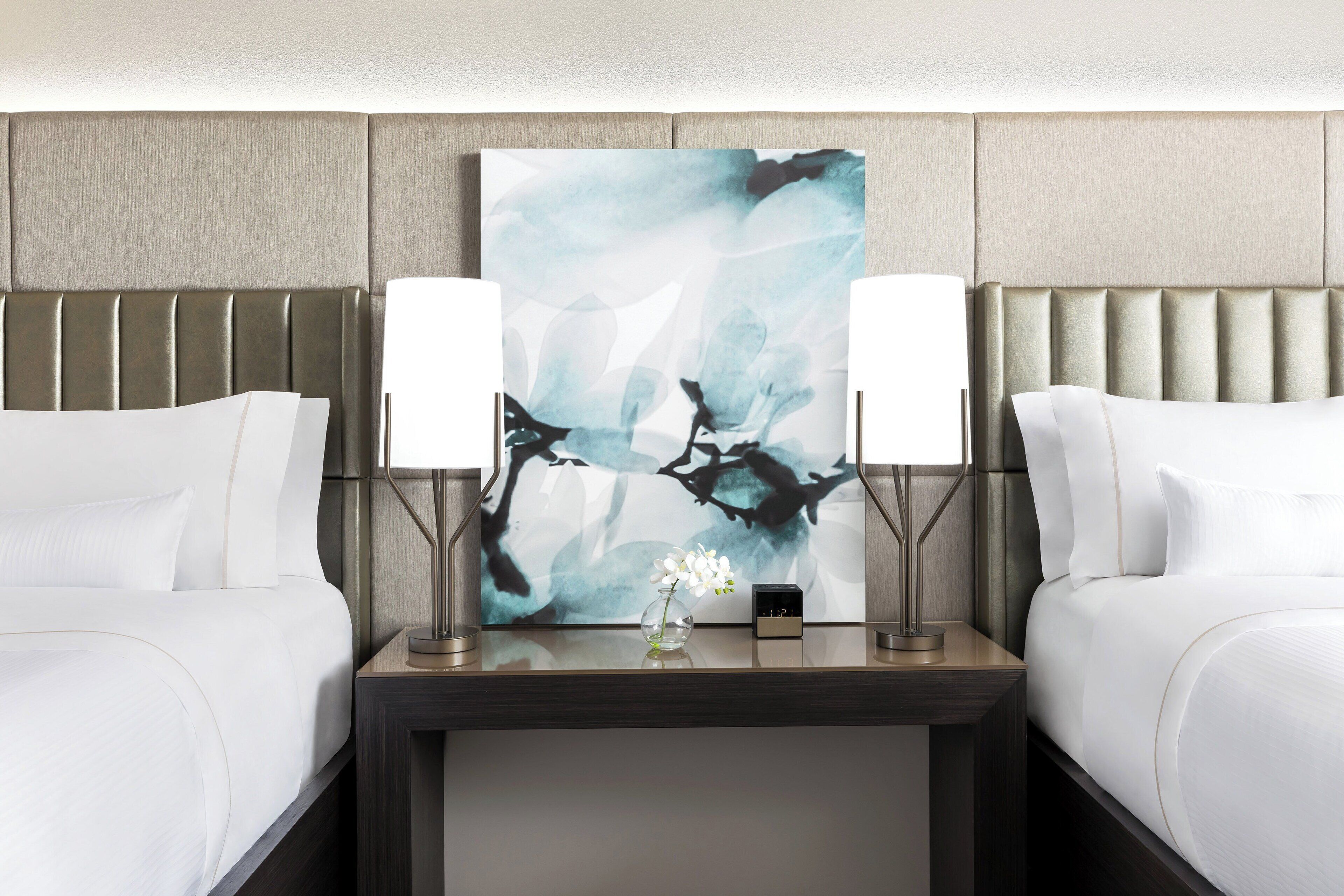 A hotel room with two beds, a nightstand with two lamps, a clock, and a painting above the nightstand.
