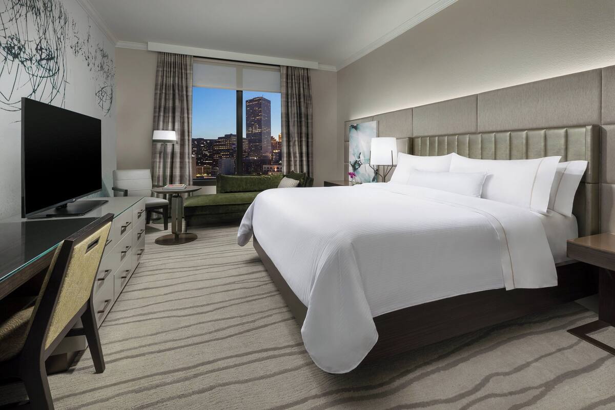 A hotel room with a large bed, a desk with a flat-screen TV, a chair, and a view of a cityscape through the window.