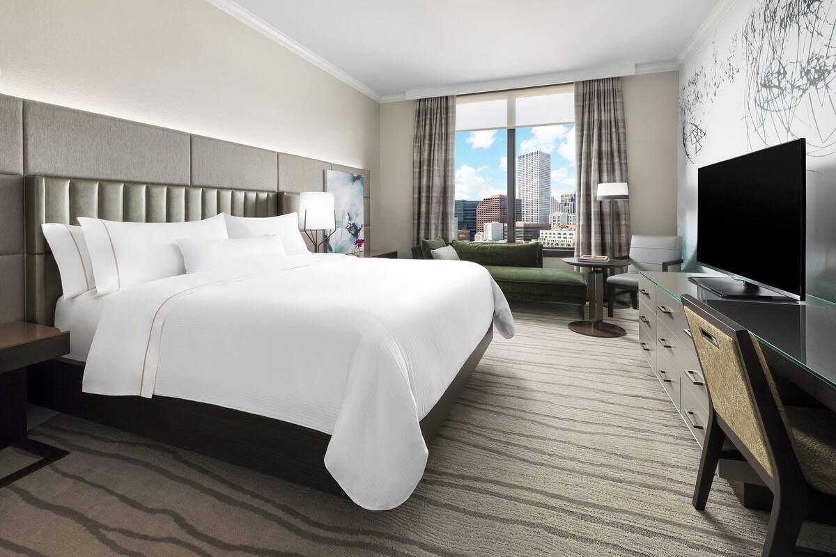 A modern hotel room with a large bed, a flat-screen TV, a desk with a chair, and a view of the city through large windows.