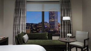 A hotel room with a bed, a green sofa, a grey armchair, a small round table with a vase of flowers, and a view of a cityscape at night. - The Westin New Orleans (New Orleans)