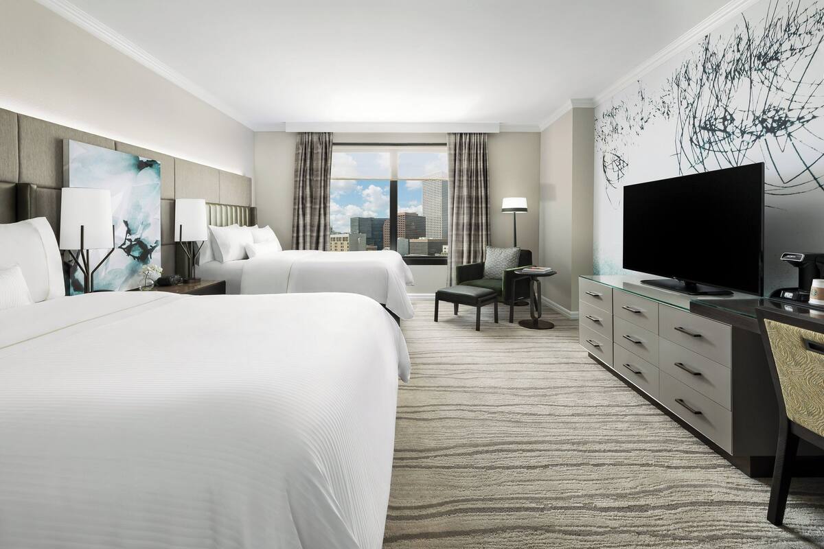 A hotel room with two beds, a desk, a television, and a large window with a city view.