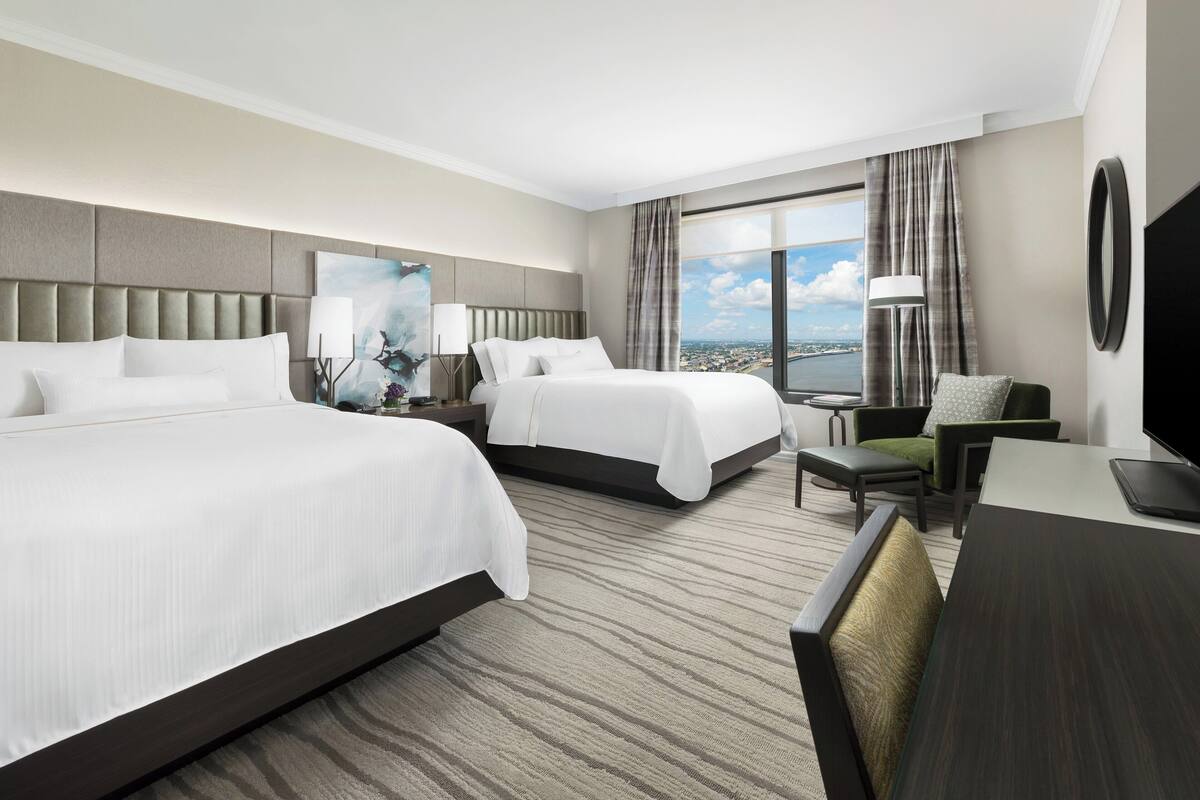 A hotel room with two beds, a desk, a chair, a TV, and a large window with a city view.