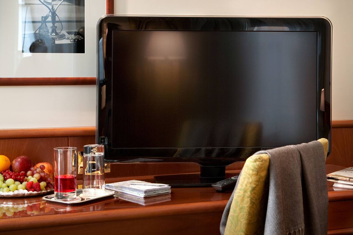 Hotellihuone, jossa on suuri litteänäyttöinen televisio, puinen työpöytä, lasi punaista juomaa, lautanen täynnä hedelmiä ja kaukosäädin.